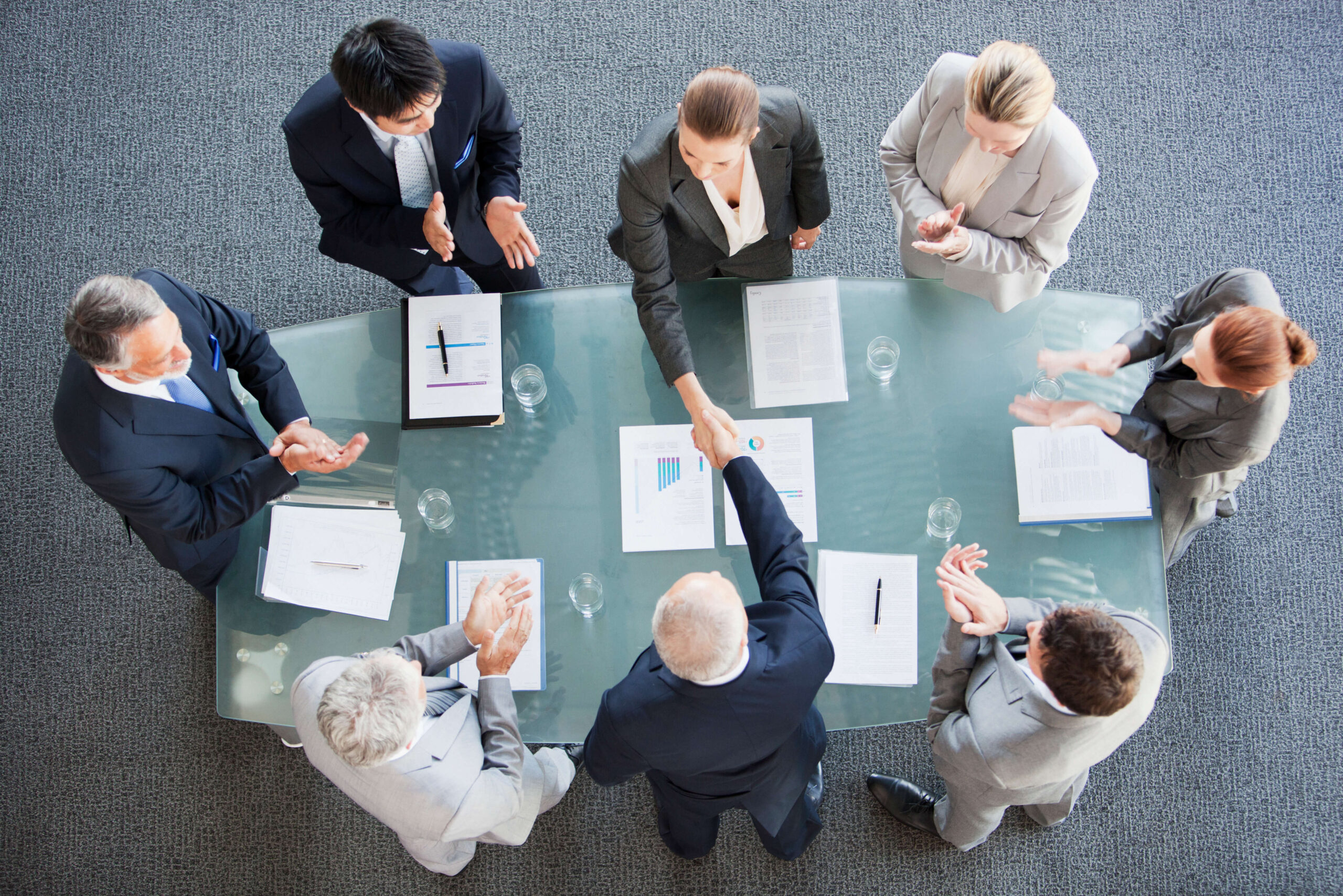 Eight business people standing around a table with documents shaking hands.