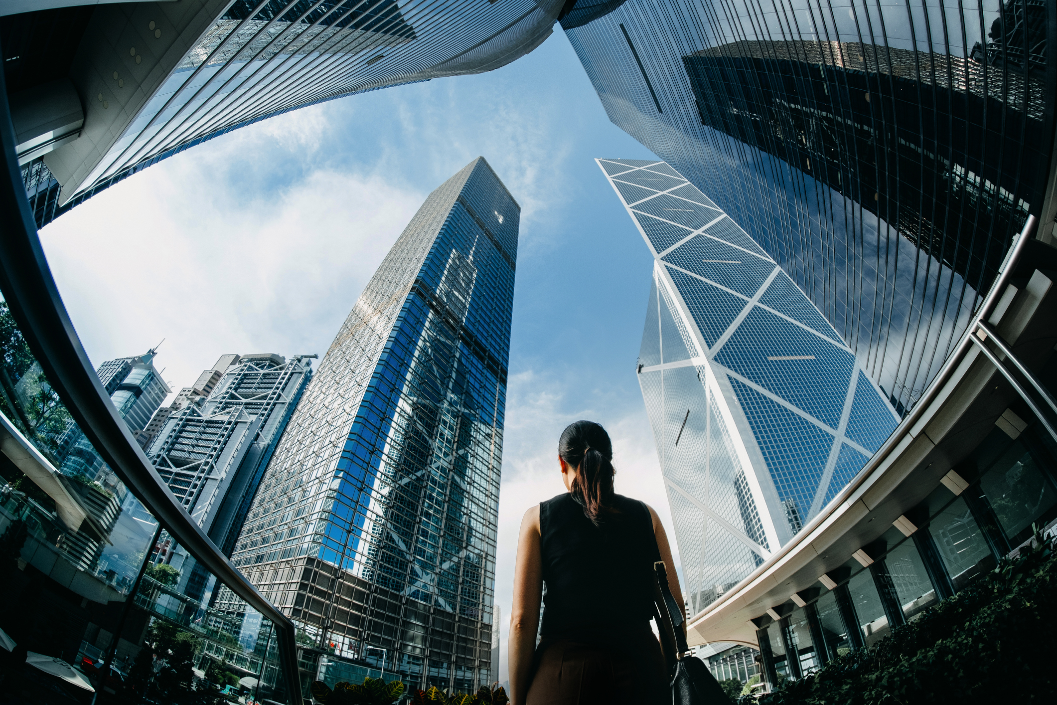 Rear view of professional young businesswoman standing against contemporary financial skyscrapers in downtown financial district and looking up into sky with positive emotion