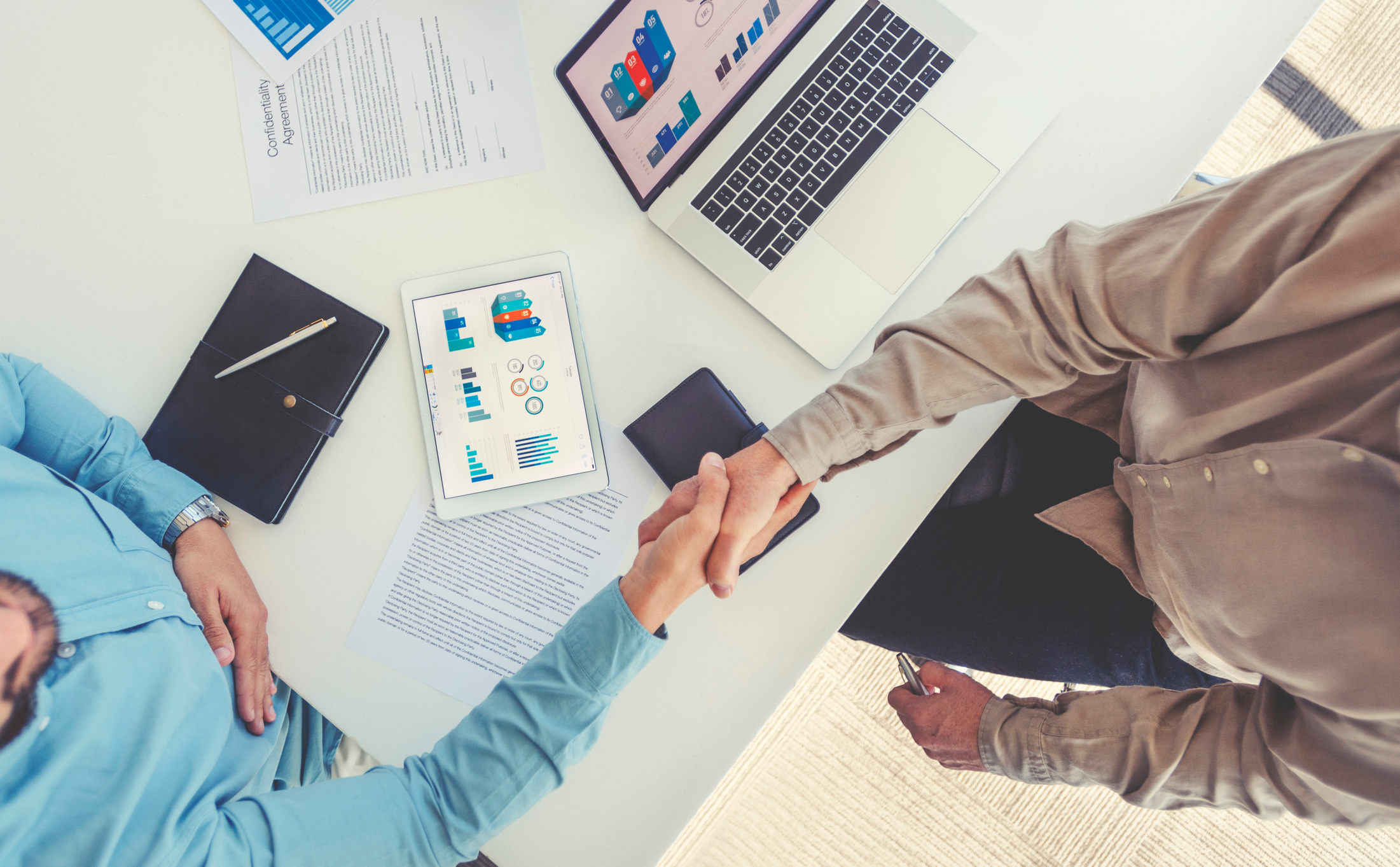 Two people shaking hand over a desk with charts and a laptop, shot from above