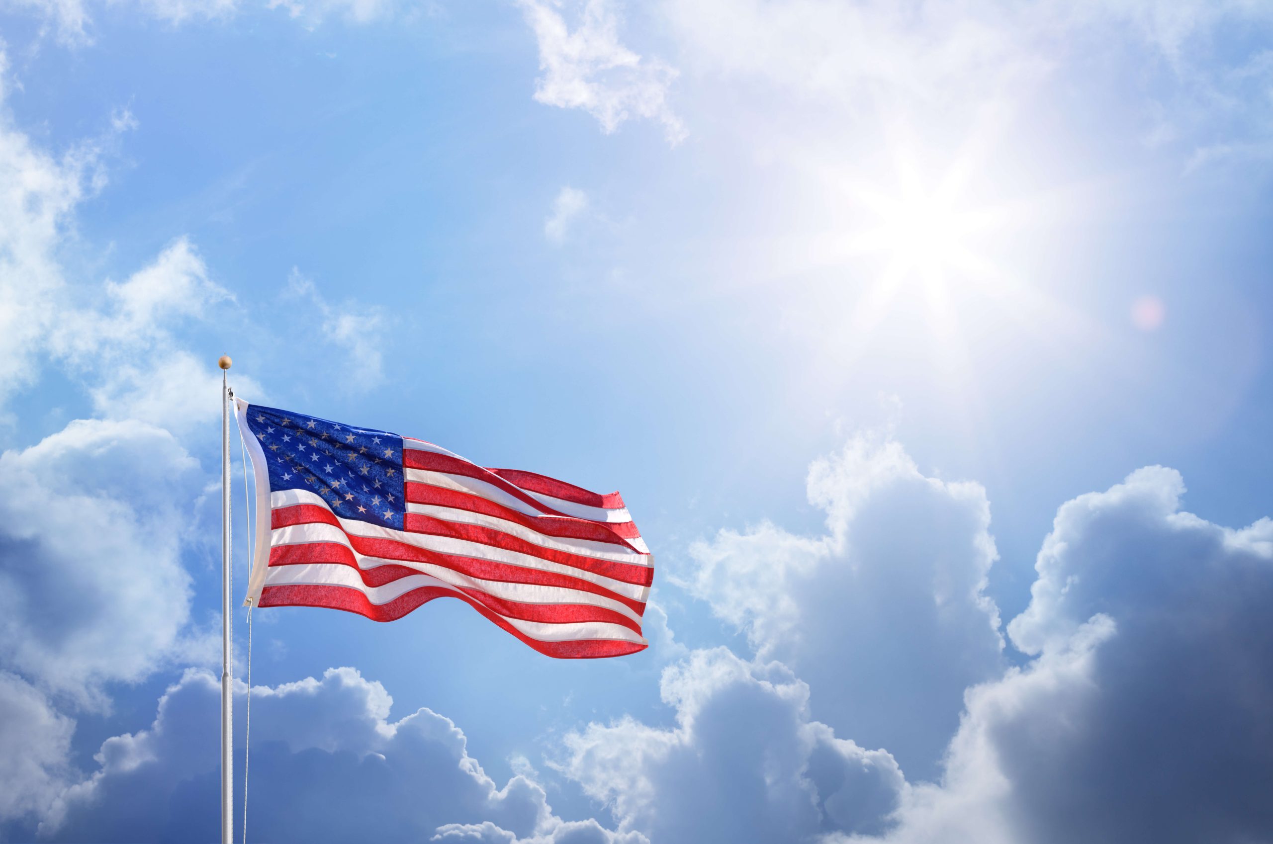 United States flag waving from a flagpole in front of a partially cloudy sky with the sun out