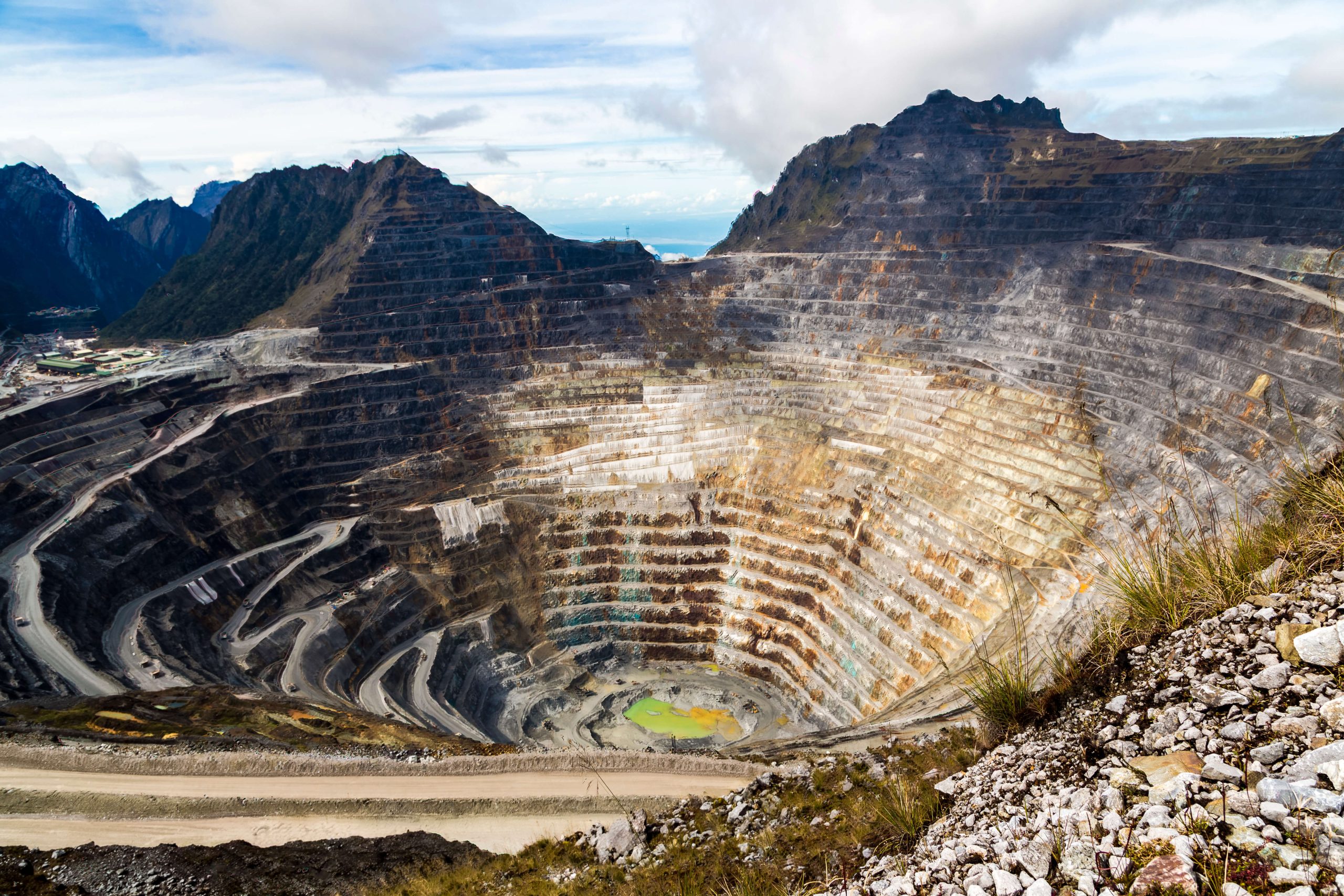 A large open-pit mine with terraced layers and a road spiraling down to the bottom, set against a backdrop of mountains and a cloudy sky.