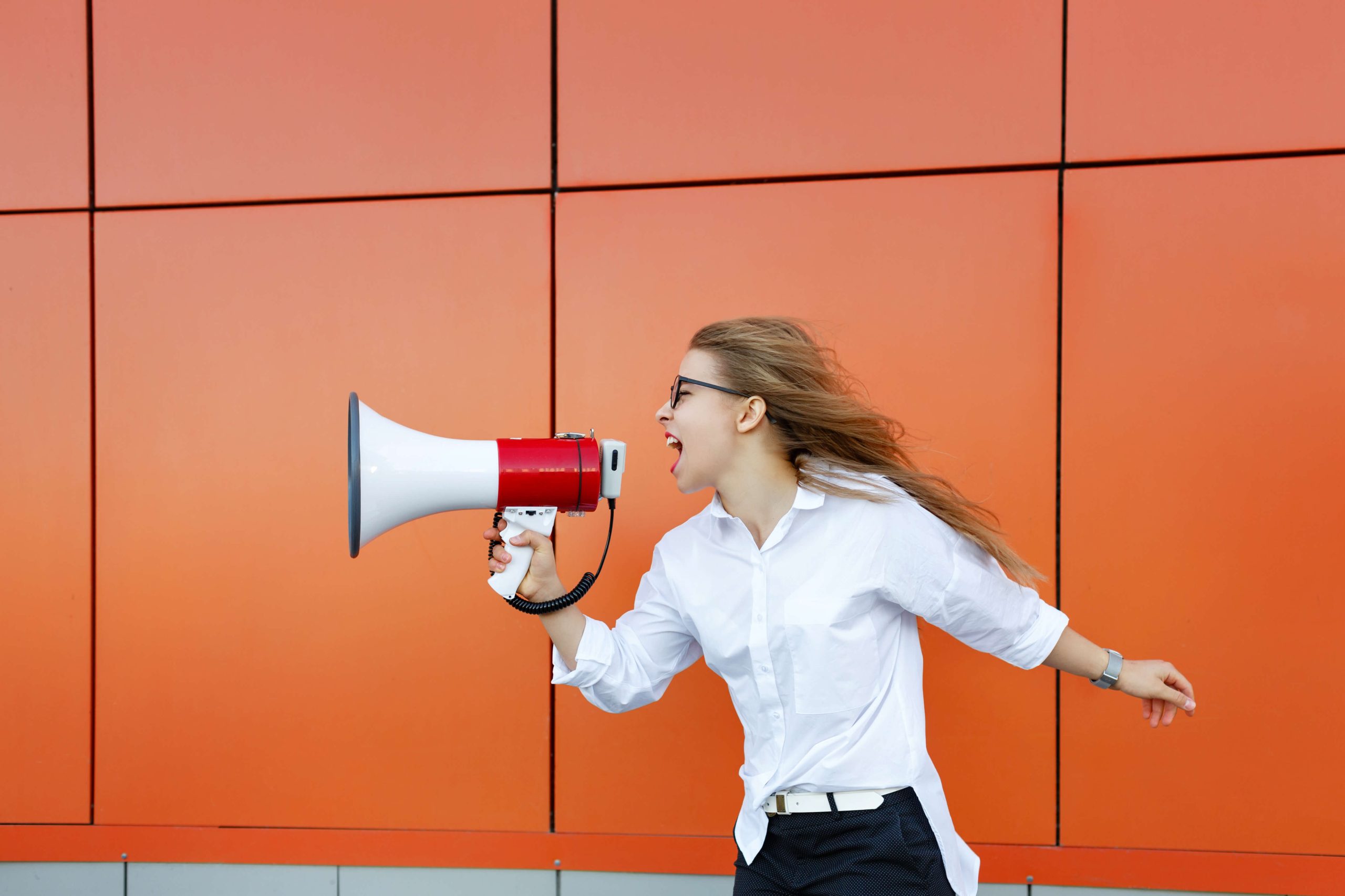 profile of windswept woman screaming into megaphone, orange plate wall behind